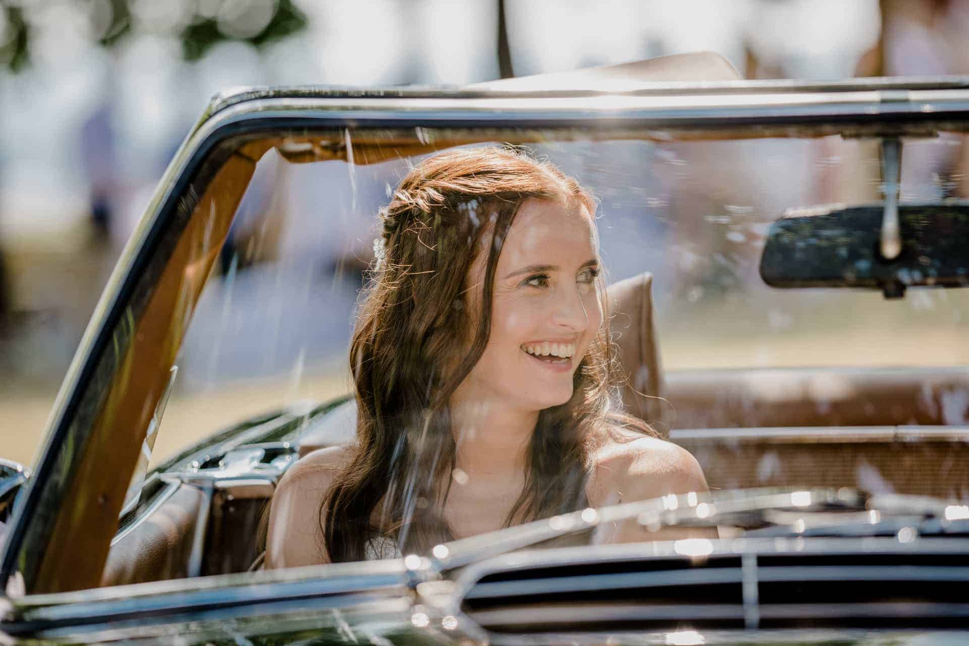 Die Braut sitzt lachend im offenen Hochzeitswagen.