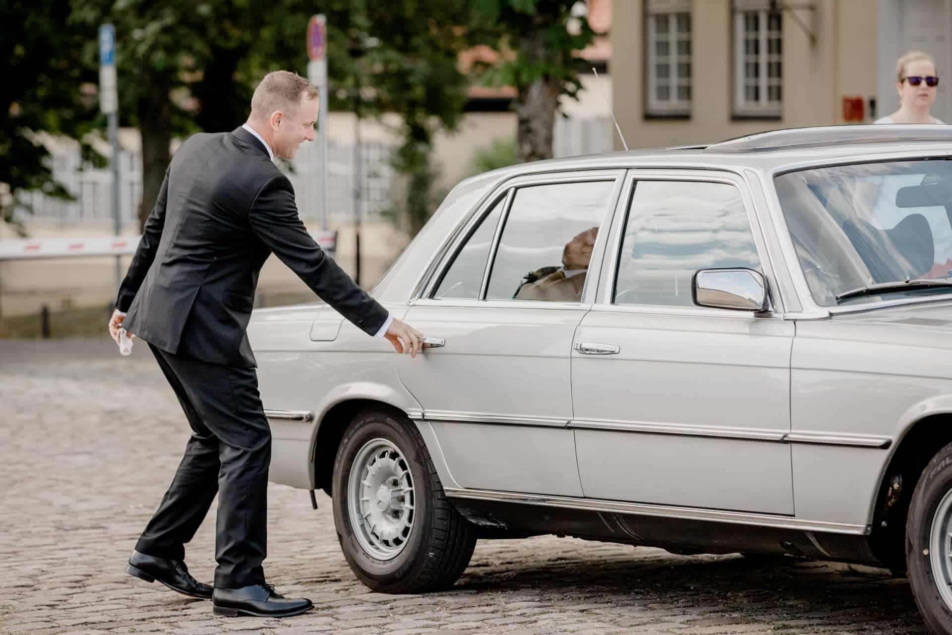 Bräutigam öffnet die Tür des weißen Hochzeitswagens