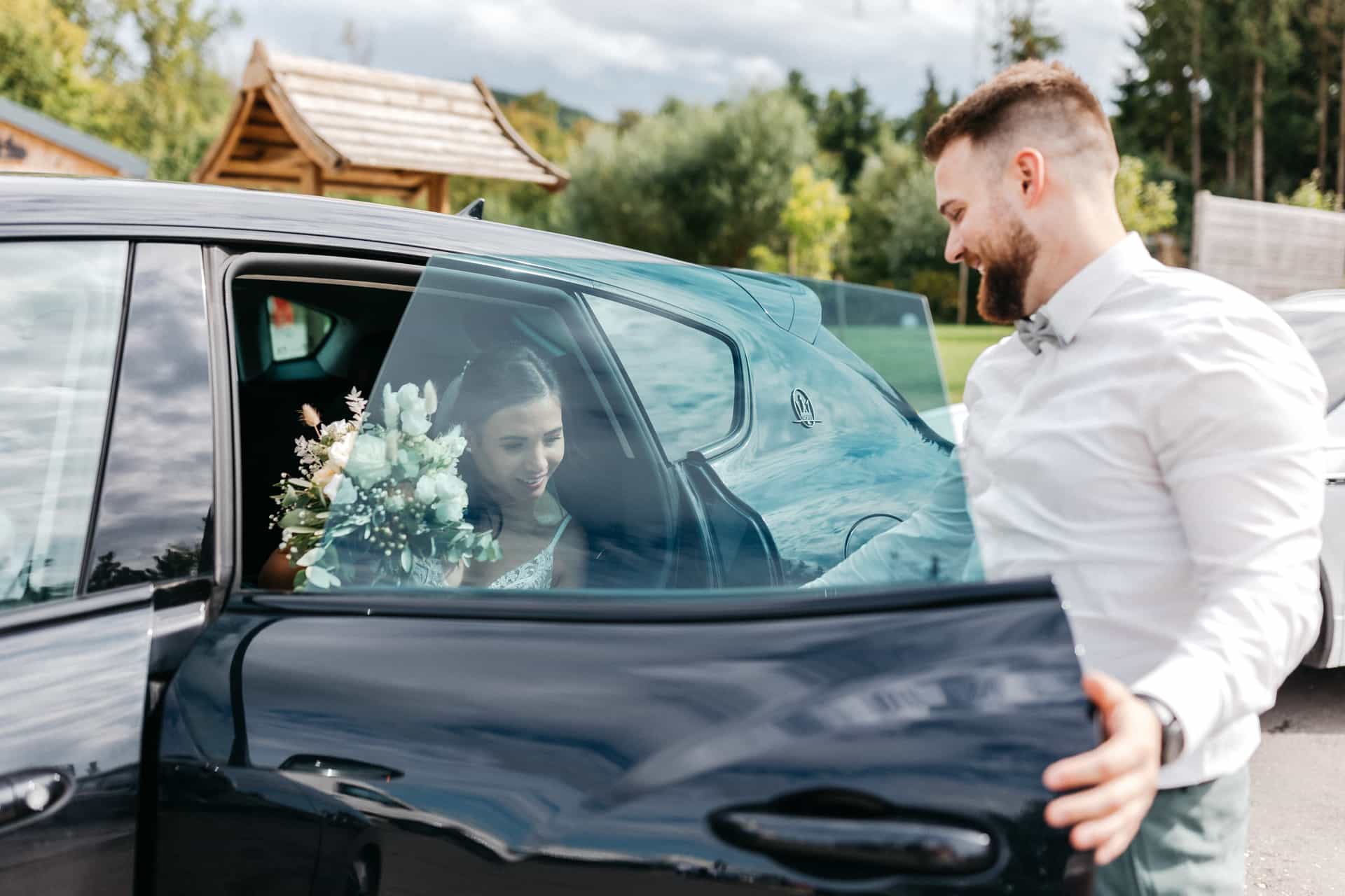 Ein Mann hält die Autotür für eine Frau mit Blumenstrauß auf.
