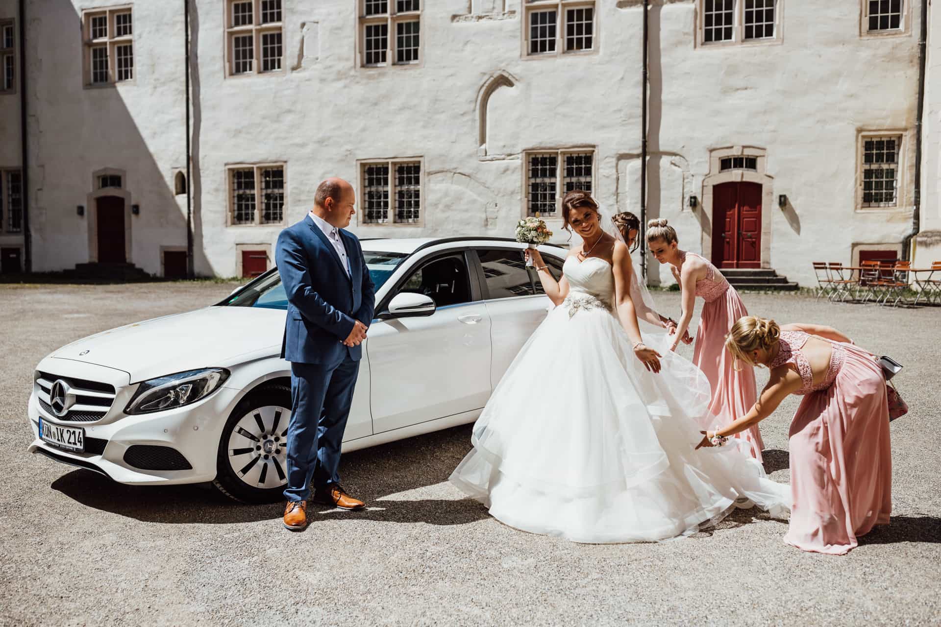 Braut und Begleiterinnen neben dem weißen Hochzeitswagen
